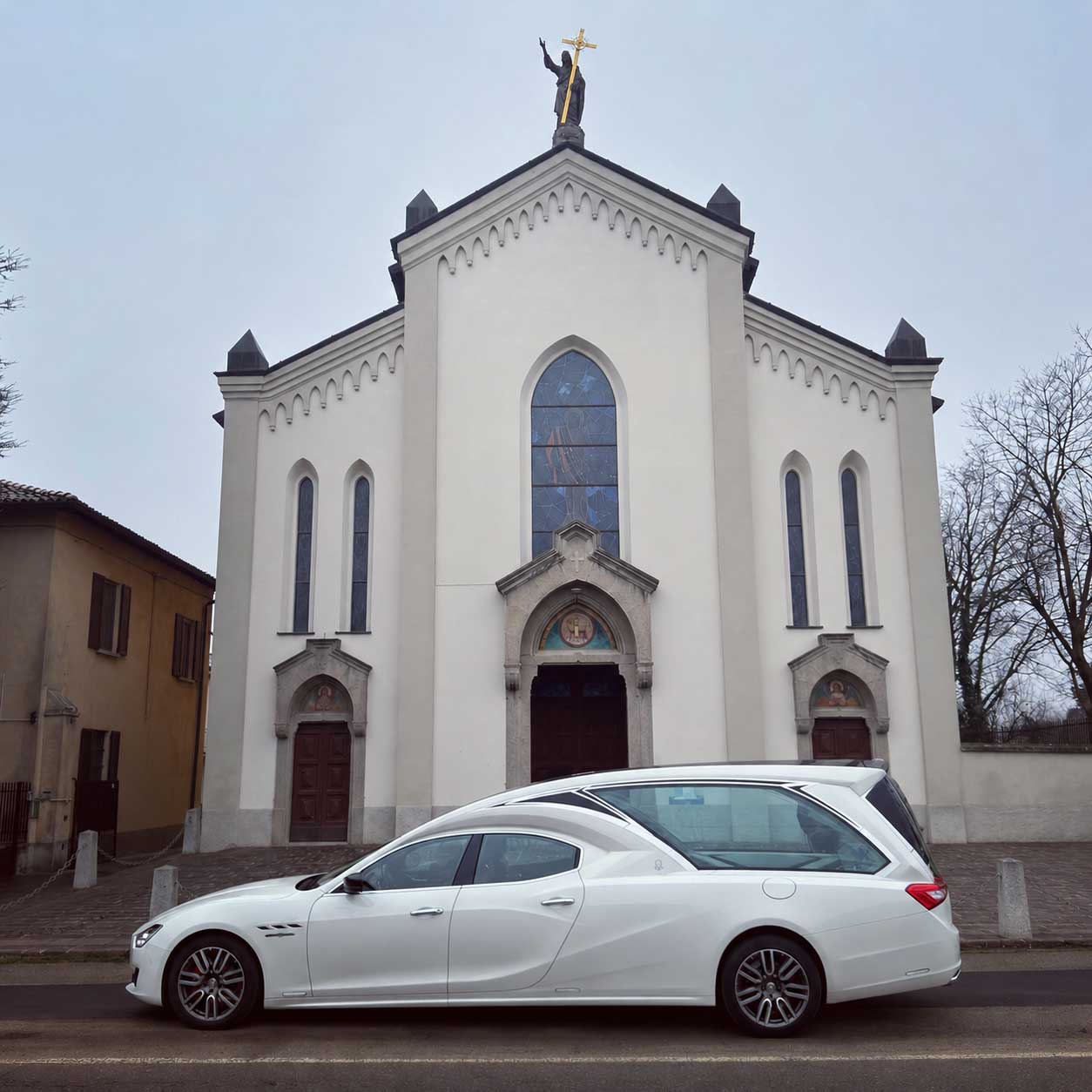 Carro funebre bianco parcheggiato davanti a una chiesa con facciata chiara, vetrate colorate e statua dorata in cima