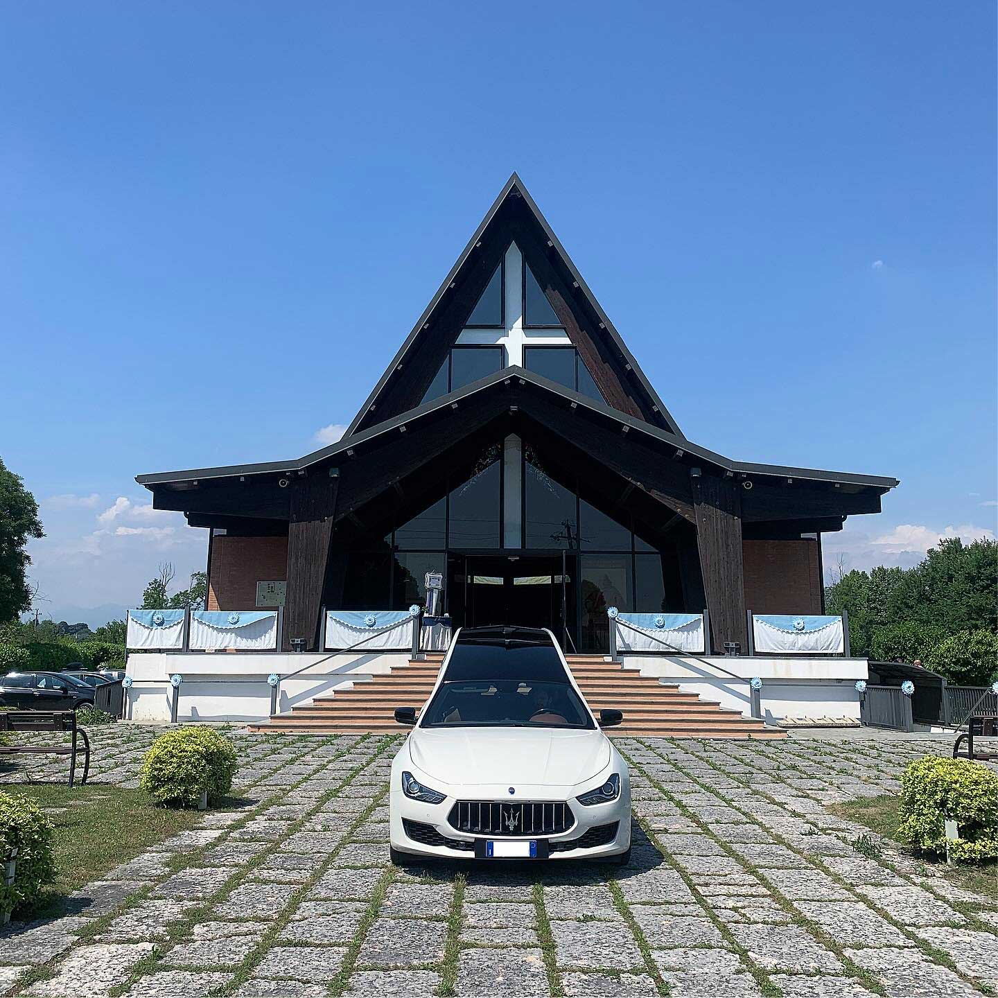 Maserati bianca davanti a una chiesa moderna con tetto a punta in legno scuro, croce vetrata e scalinata, su selciato