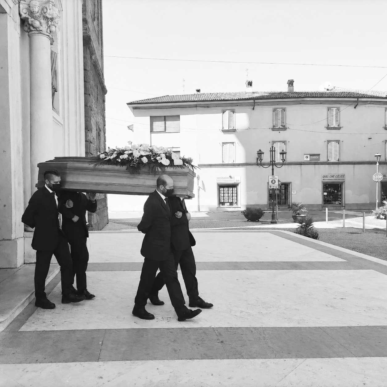 Quattro necrofori in abito scuro e mascherina portano a spalla una bara di legno con fiori bianchi davanti a una chiesa