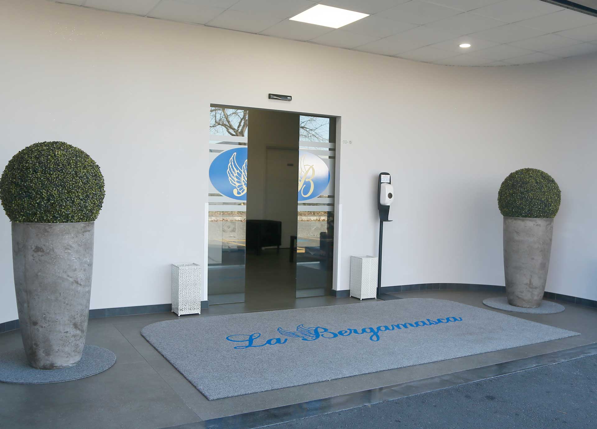 Ingresso della Casa del Commiato La Bergamasca con porte a vetri, logo blu alato, tappeto con scritta e due vasi di bosso
